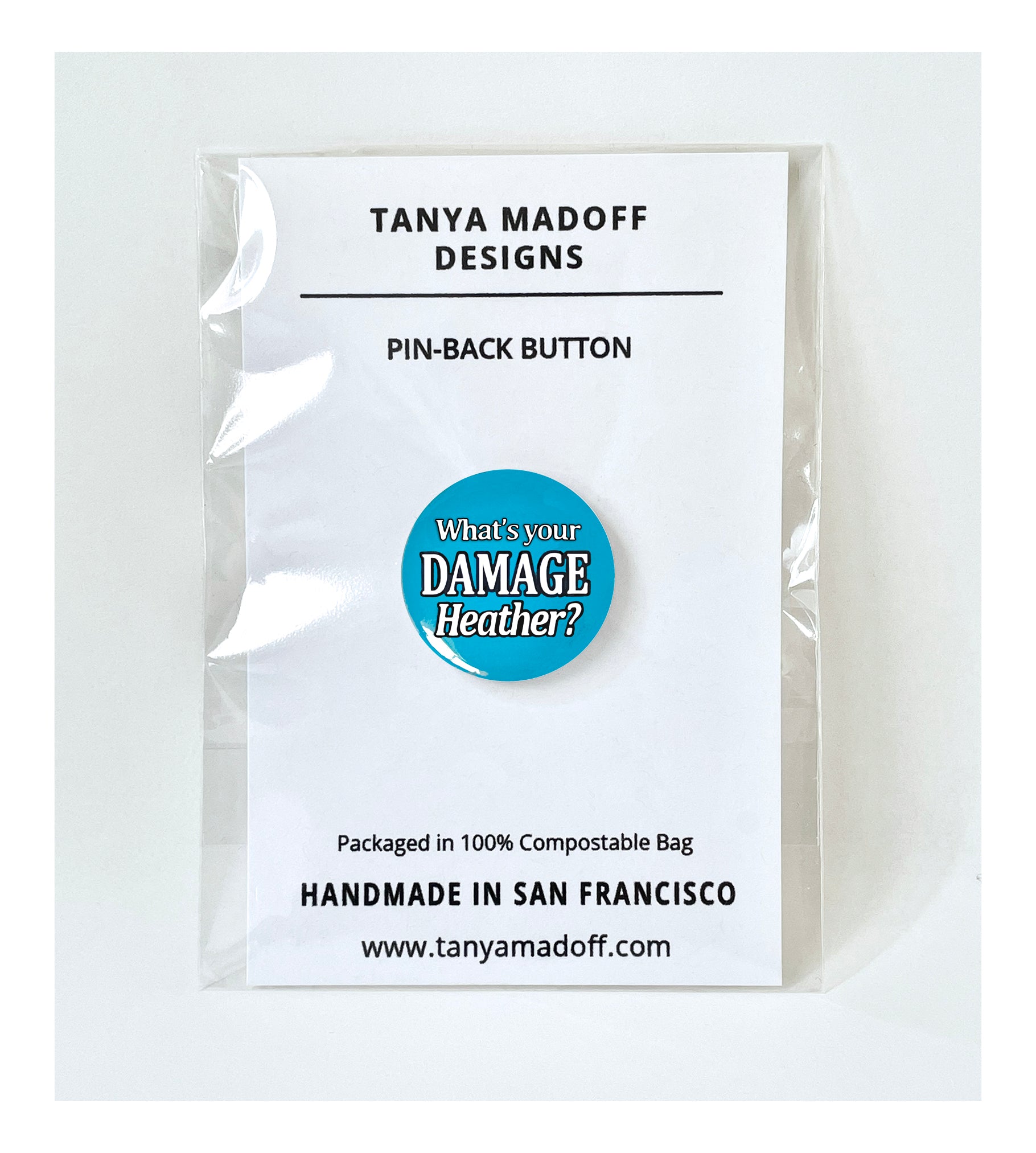 What’s Your Damage Heather? one inch pin back badge packaged on recycled paper, in 100% compostable clear glassine bag